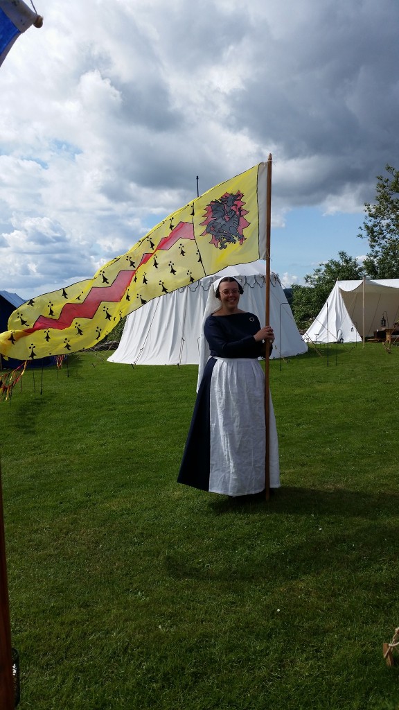 Photo of a Caucasian woman wearing a navy blue dress and a white apron holding a pole with a yellow and red heraldic banner on it, standing in front of a medieval tent.