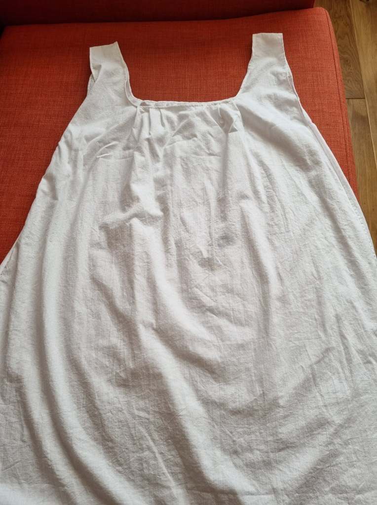 Photograph of a sleeveless white cotton shift with a square pleated neckline, laying flat on an orange sofa.